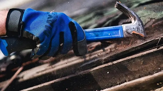hand in a blue work glove holding a blue hammer as it smashes through an old roof